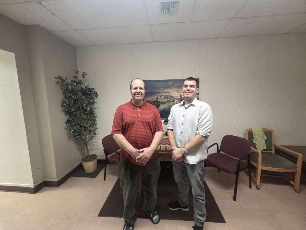 Two people stand smiling in a room with chairs, a plant, and a painting.