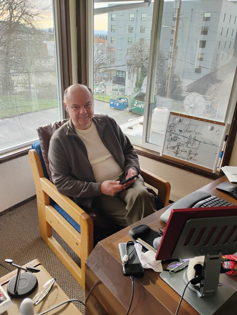 A person sitting by a window, holding a phone, with a desk nearby.