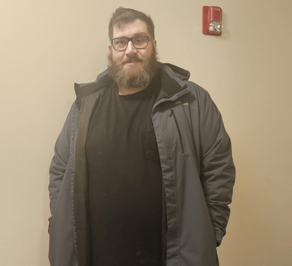 A person with a beard and glasses wearing a dark coat stands in front of a wall with a fire alarm.