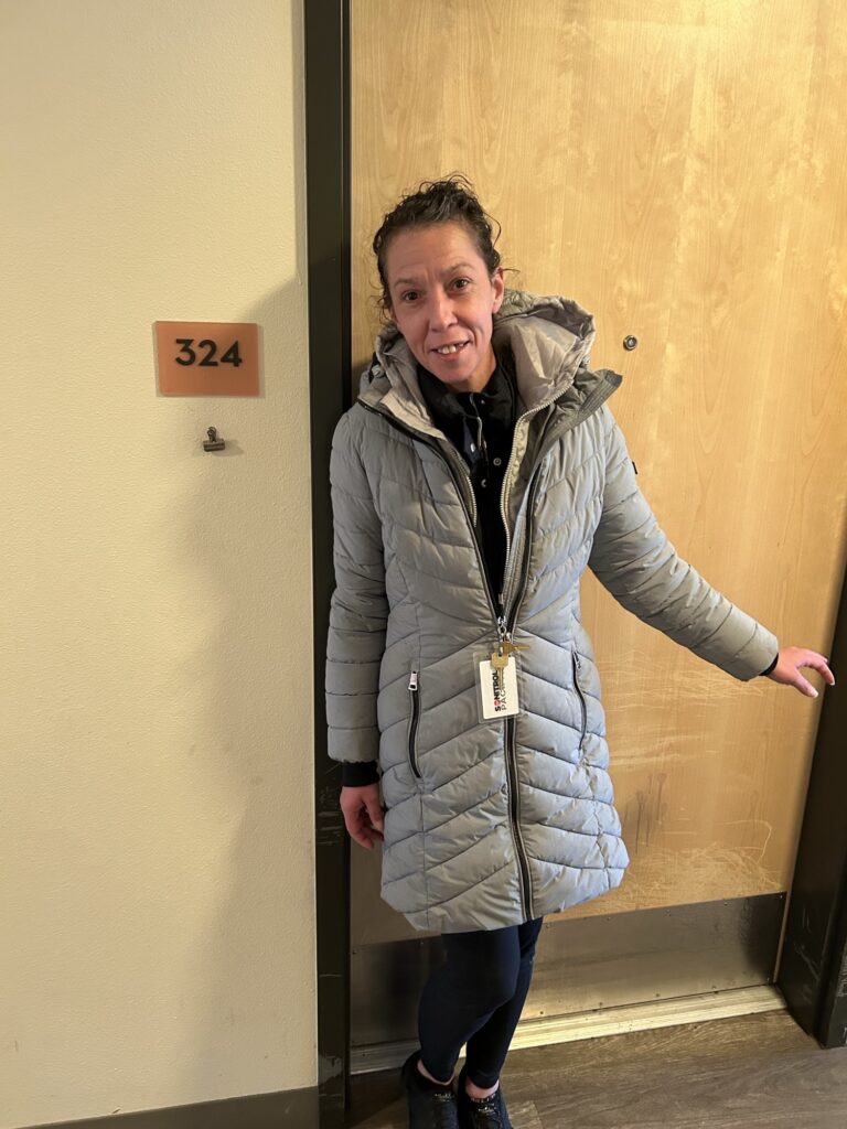A person stands smiling in a hallway near a door labeled 324.