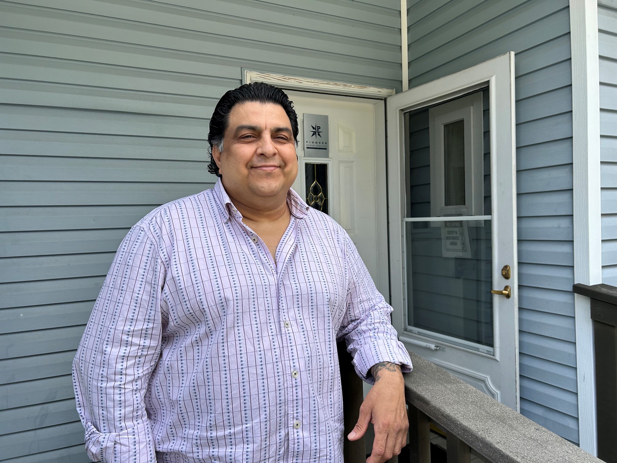 A person stands smiling on a porch, wearing a striped shirt, with a background of a blue building and door.